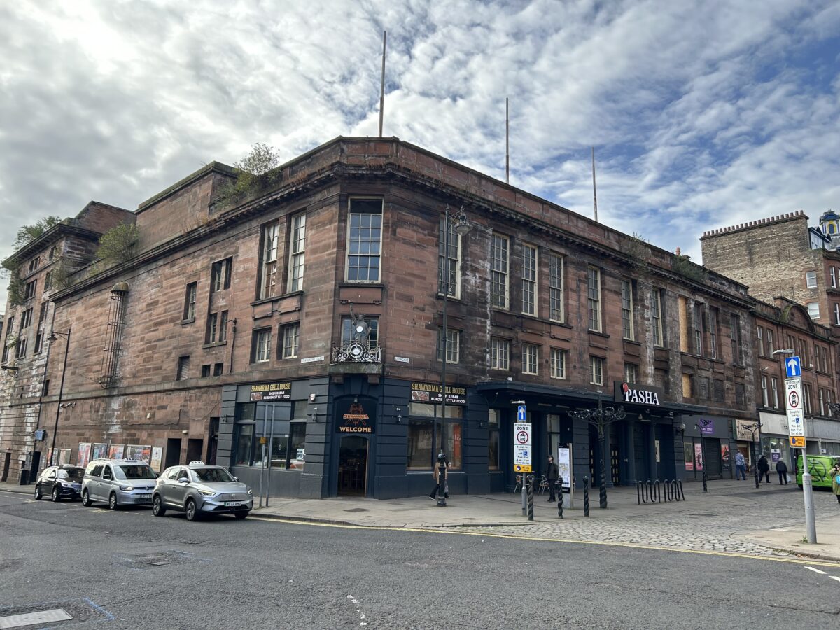 Street-corner view of a large red sandstone former theatre building with weathered façades, upper-floor windows, and modern restaurant and nightclub frontages at ground level under a cloudy sky.