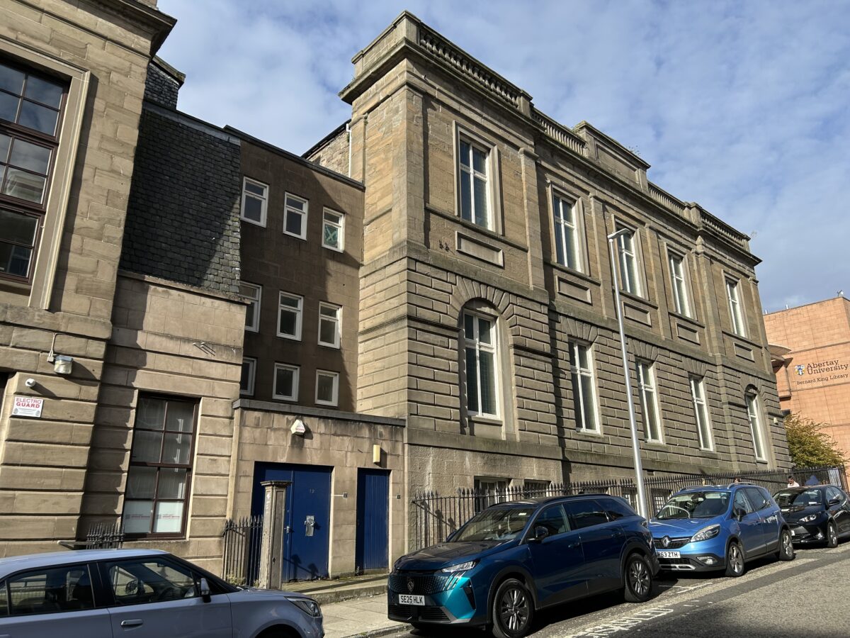 Street view of a large historic sandstone building with classical architectural detailing, tall sash windows, and a rusticated ground floor. Several cars are parked along the road in front of the building, which is enclosed by black iron railings. Abertay University’s Bernard King Library is visible in the background under a partly cloudy sky.