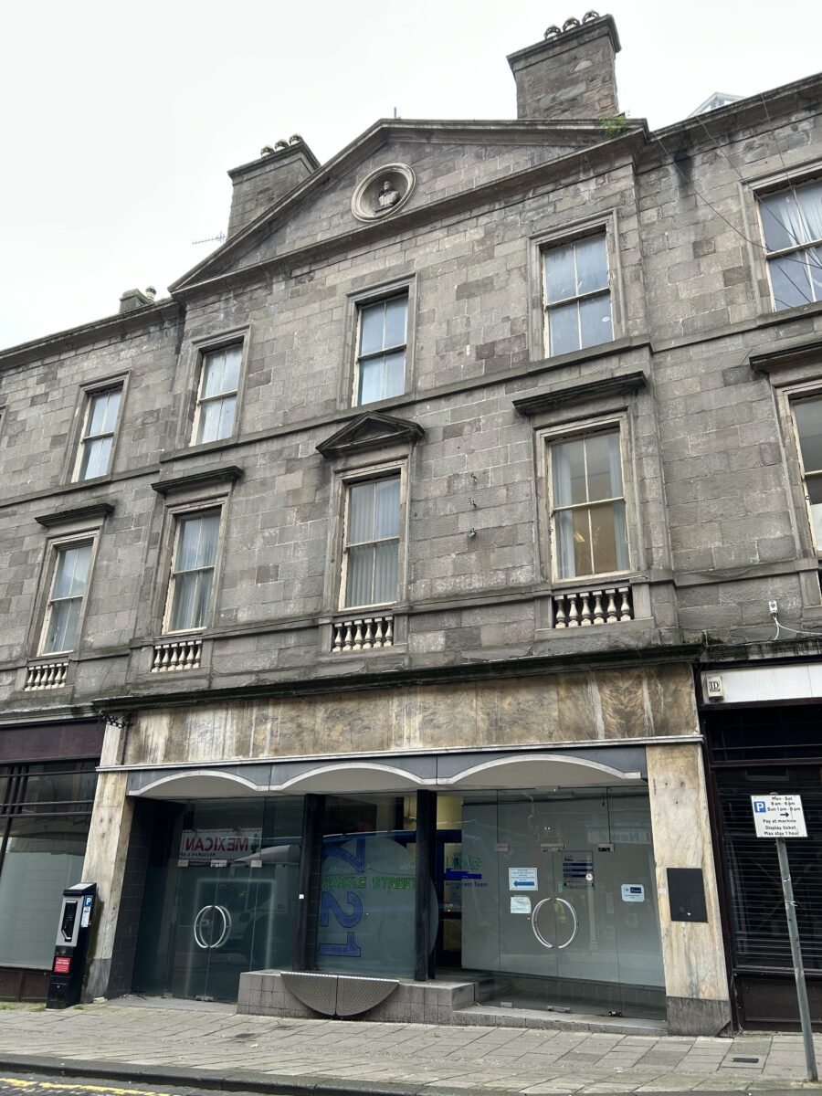 View looking up at a grey stone historic building façade, showing sash windows, a small balustraded window balcony, and a triangular pediment.