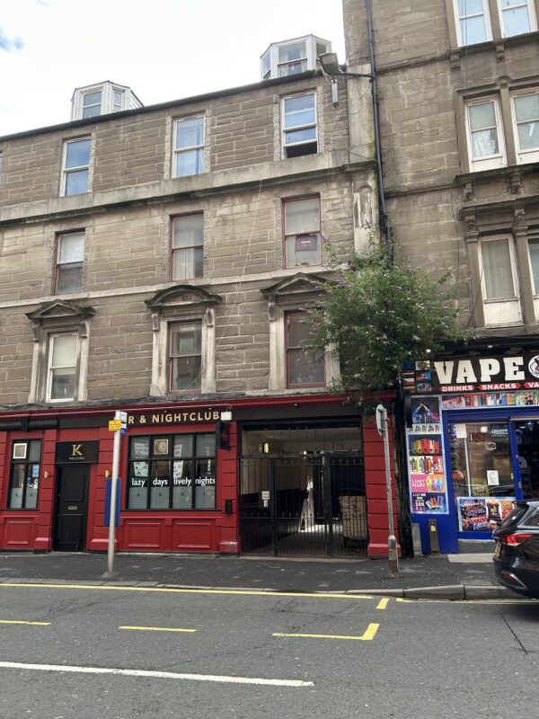 Street view of a historic stone tenement building with commercial premises at ground level. The three-storey façade features sash windows, decorative stonework, and dormer windows in the roof, with a narrow passageway leading into the building's interior.