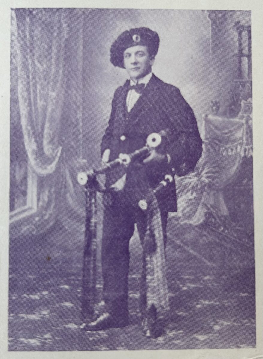 Studio portrait of a young person wearing a dark suit, bow tie and beret-style hat, standing beside a bicycle and holding bagpipes, posed against a painted photographic backdrop with drapery and a chair.
