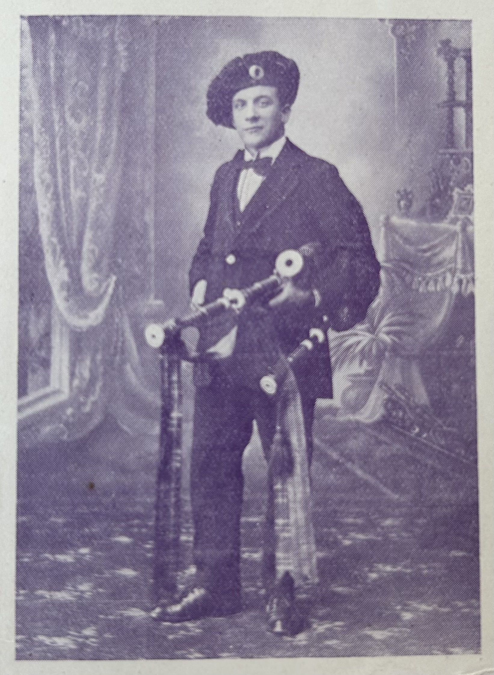 Studio portrait of a young person wearing a dark suit, bow tie and beret-style hat, standing beside a bicycle and holding bagpipes, posed against a painted photographic backdrop with drapery and a chair.